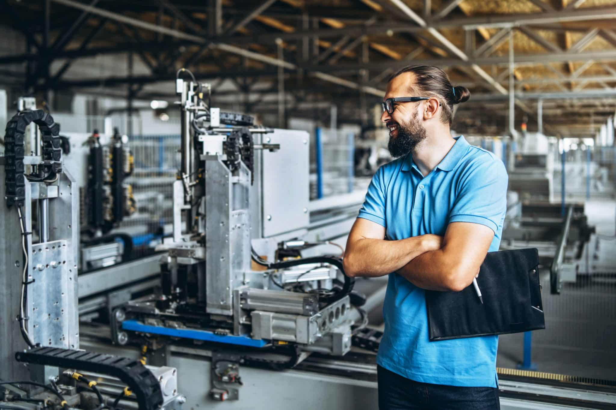 Junger Ingenieurmanager mit Bart überprüft Fertigung, Arbeitsplatz und Maschinen in einer großen Fabrik.