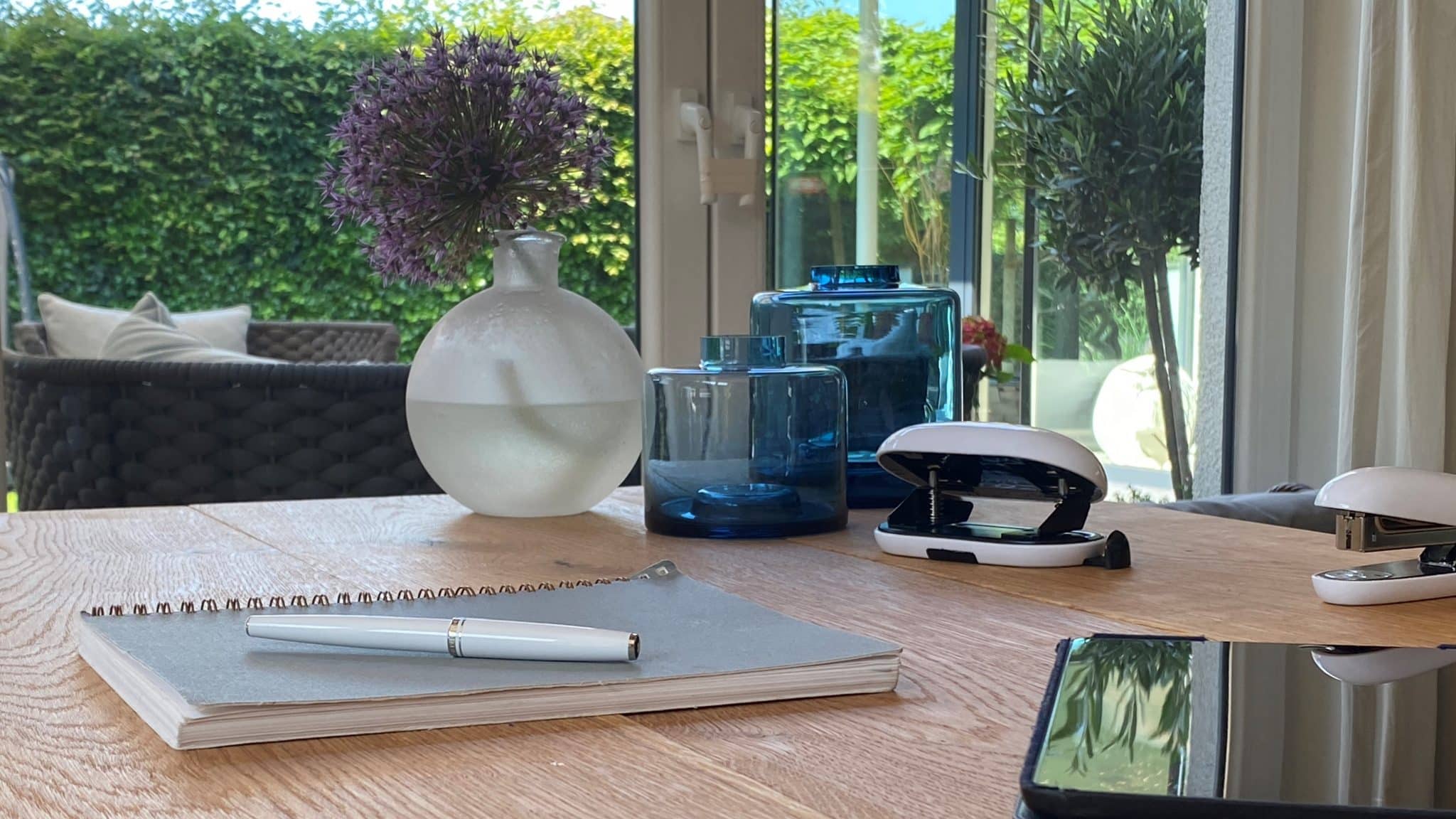 Ein heller Homeoffice-Platz wird von einem Notizbuch, einer Vase und blauen Glasgefäßen begleitet.