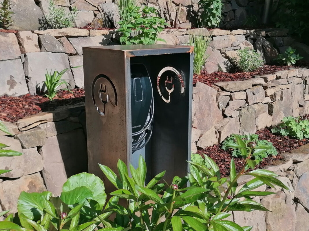 Cortenstahl-Wallbox mit Ladestation vor bepflanzter Natursteinmauer im Außenbereich der privaten Auffahrt integriert.