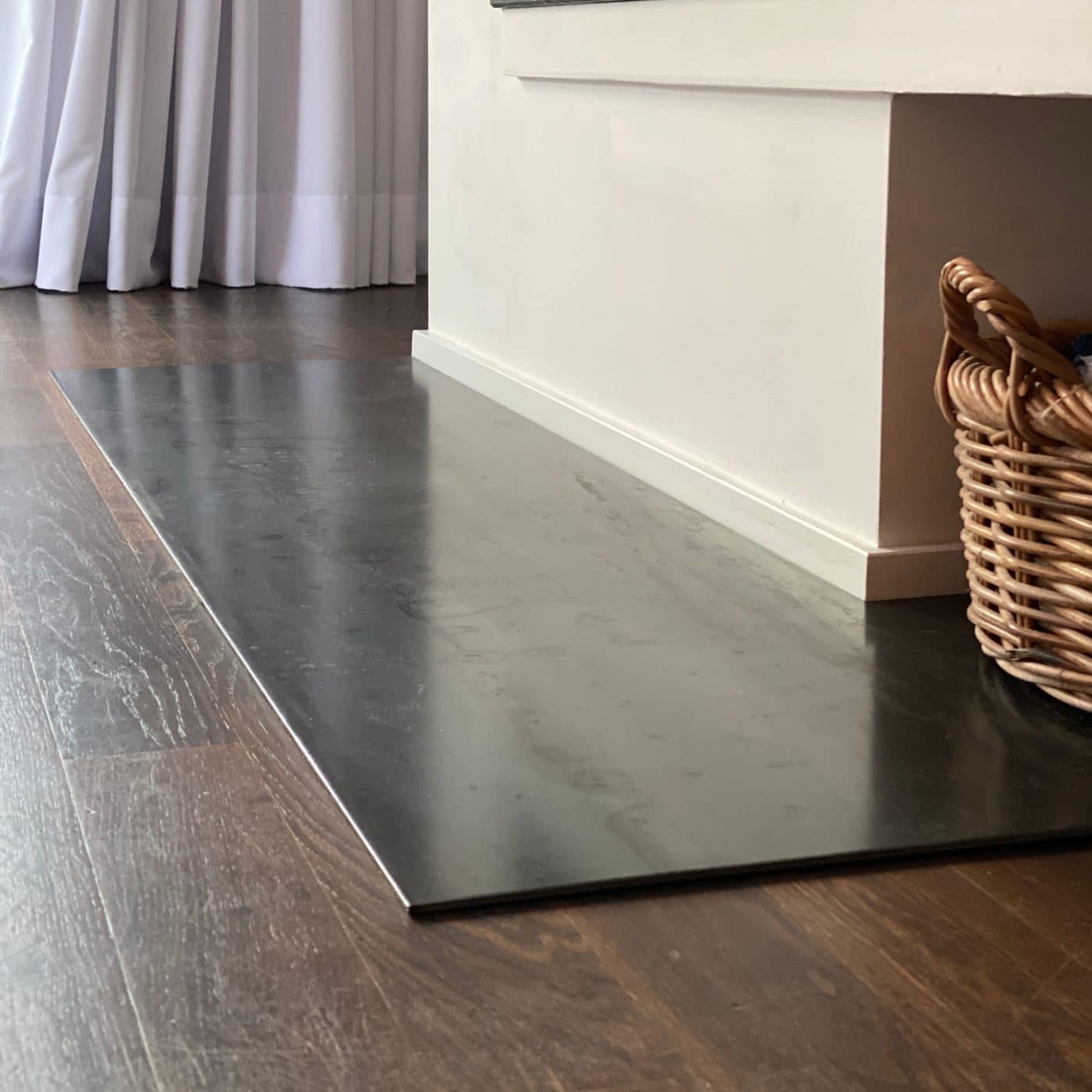 Funkenschutzplatte aus Metall vor Kamin auf dunklem Holzboden mit einem Holzkorb daneben.