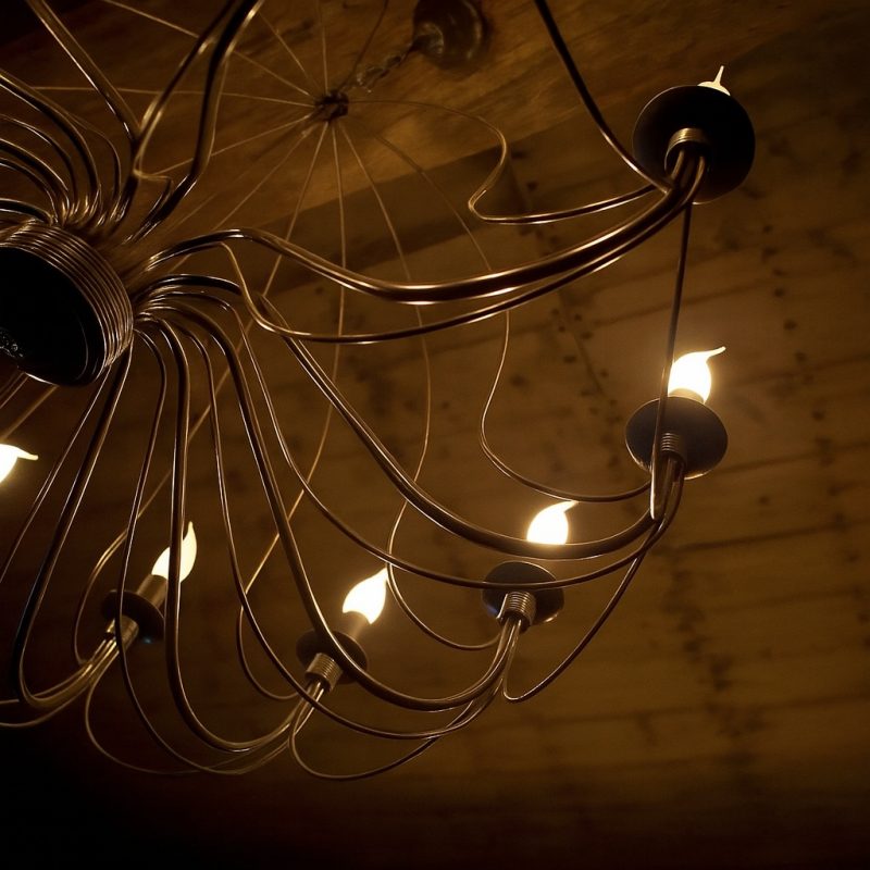 Detailaufnahme eines modernen Kronleuchters mit geschwungenen Metallarmen und warmem Licht vor Holzdecke.