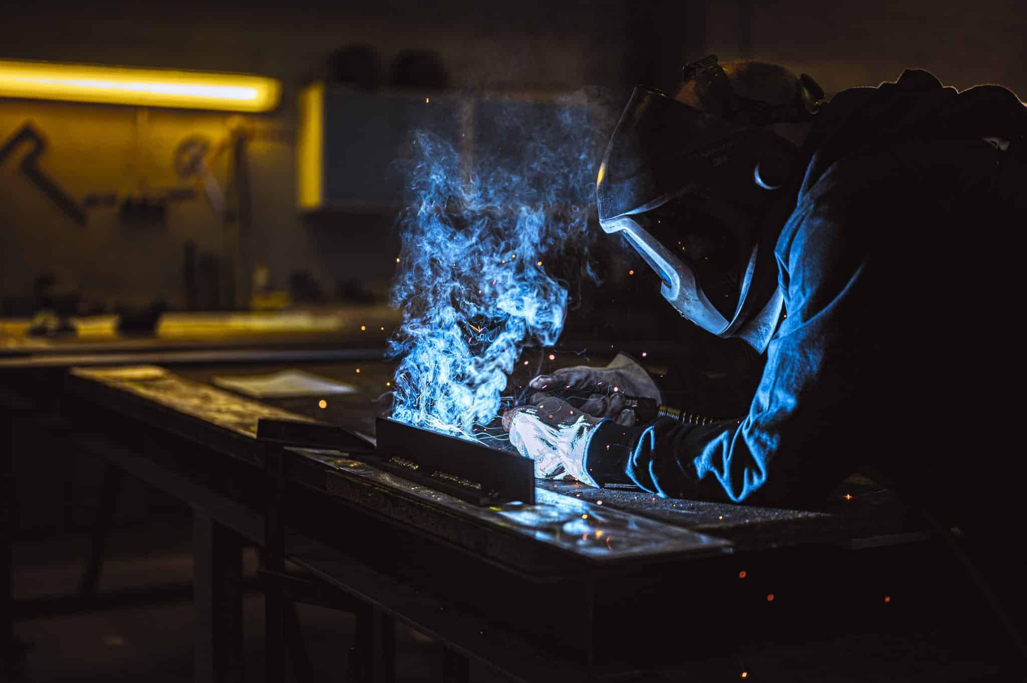 Schweißer bei der Metallbearbeitung mit blauem Licht und Funken in dunkler Werkstatt.