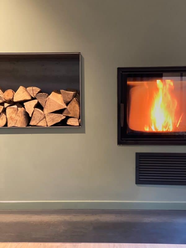 Brennender Kamin mit Holzlager und dunkler Funkenschutzplatte vor dem Ofen.