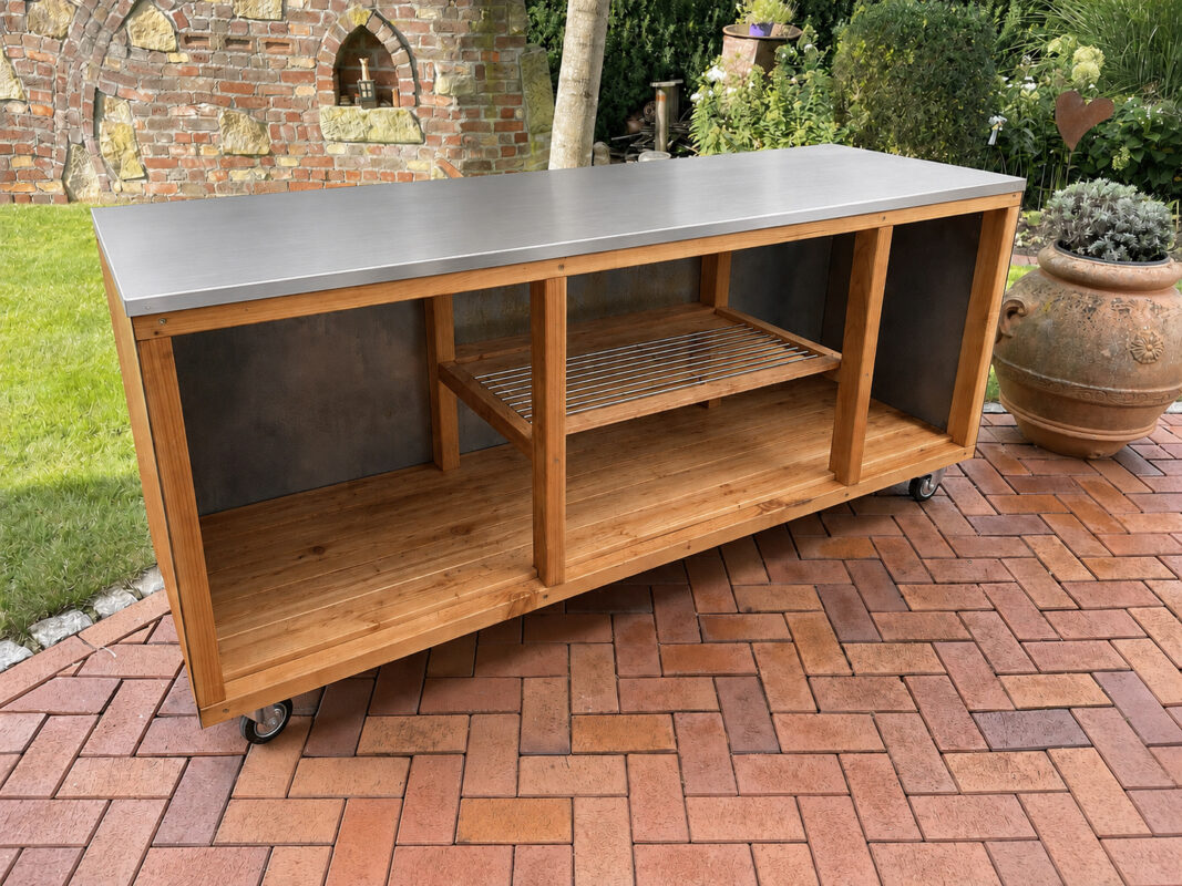 Mobile Outdoorküche mit Edelstahl-Arbeitsplatte, Holzrahmen und offenen Ablagefächern auf einer Gartenterrasse im Außenbereich.