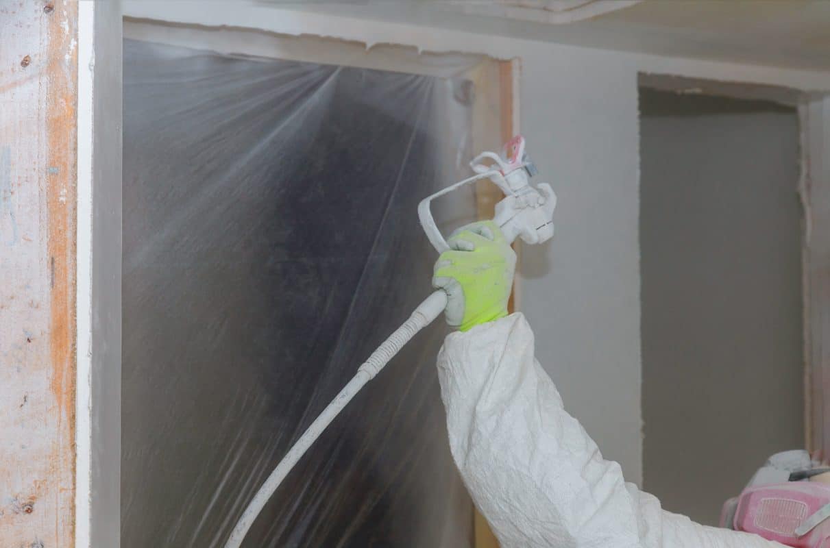 Person sprüht Wandfarbe mit Farbsprühgerät auf.