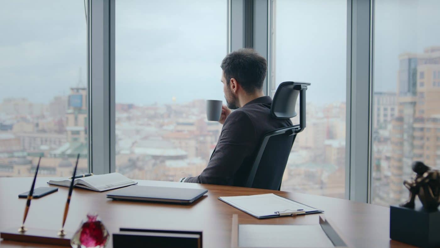 Mann im Büro schaut mit Kaffeetasse aus dem Fenster über die Stadt.